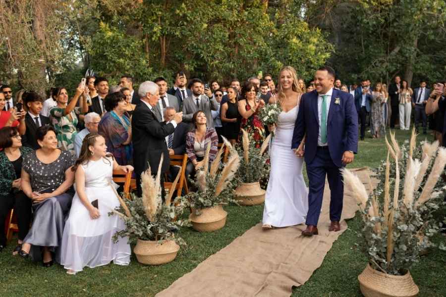 Un couple de jeunes mariés marche joyeusement le long d'une allée extérieure lors de leur cérémonie, sous le regard attendri des invités.