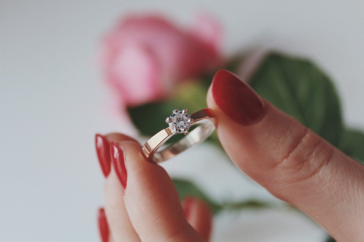 Une main aux ongles rouges présente une élégante Bague Femme Plaqué Or ornée d'une pierre scintillante devant une rose.