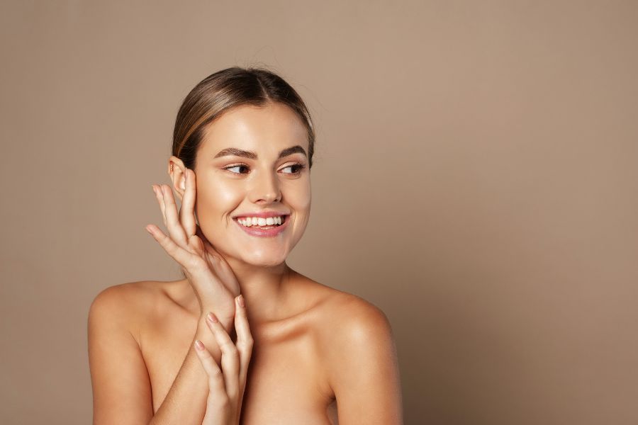Une jeune femme souriante au teint lumineux touche délicatement sa joue dans un studio au fond neutre